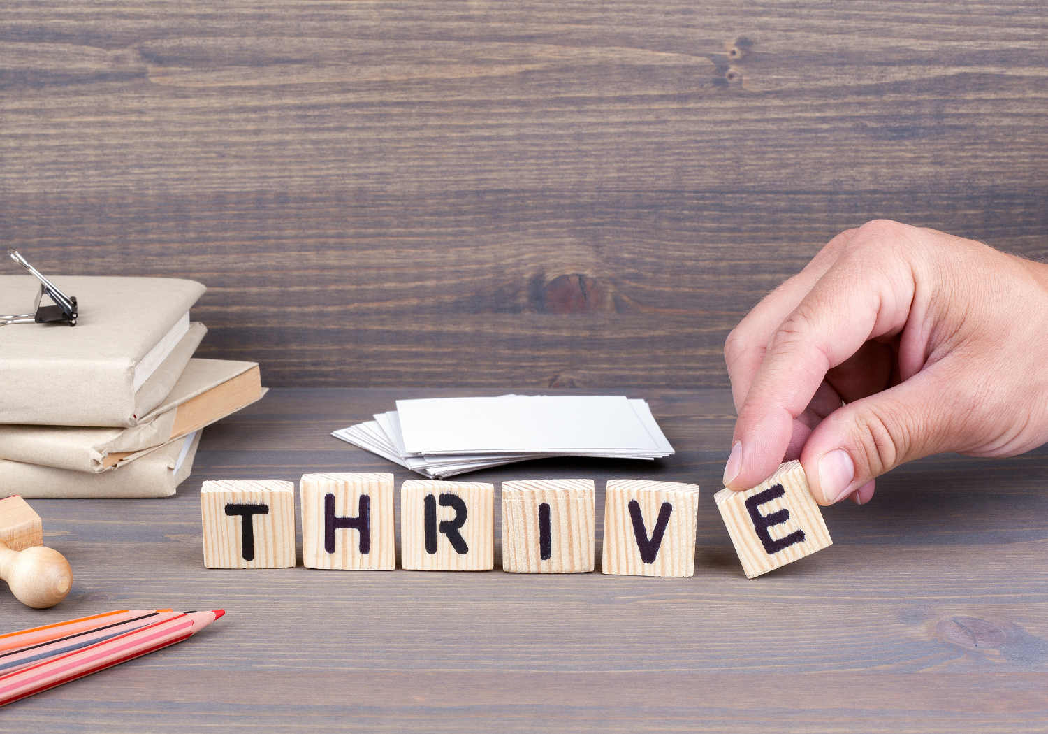 Small wooden blocks that spell the word thrive, a hand is placing the final letter, the E. 
