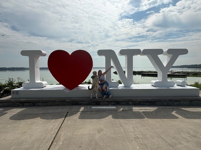 a woman poses with her dog, jethro, in front of the "I <3 NY" sign in Geneva NY